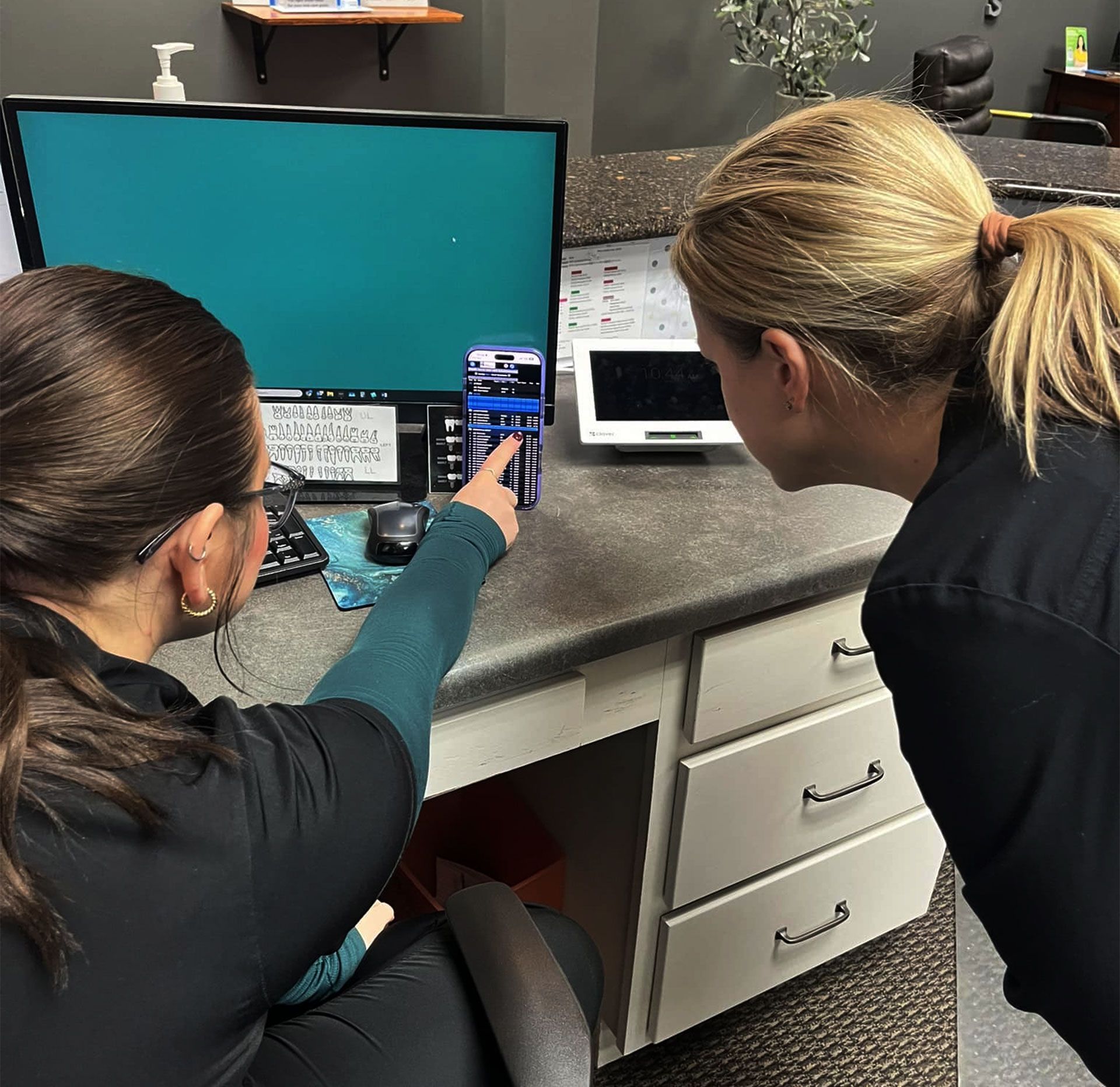 Two professionals standing at a desk, looking at a desktop computer monitor and a small handheld device.