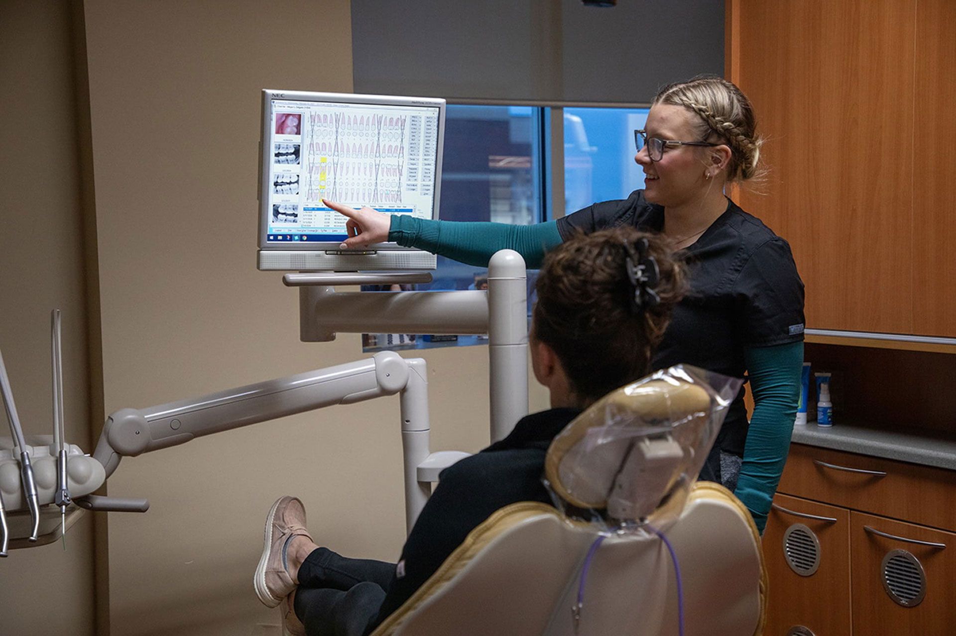 A dental professional explains dental chart information on a computer screen to a patient sitting in the dental chair.