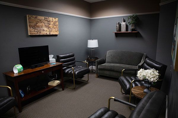 A cozy waiting room with a TV, a wooden world map, a dark gray sofa, four black leather chairs, and a small side table.