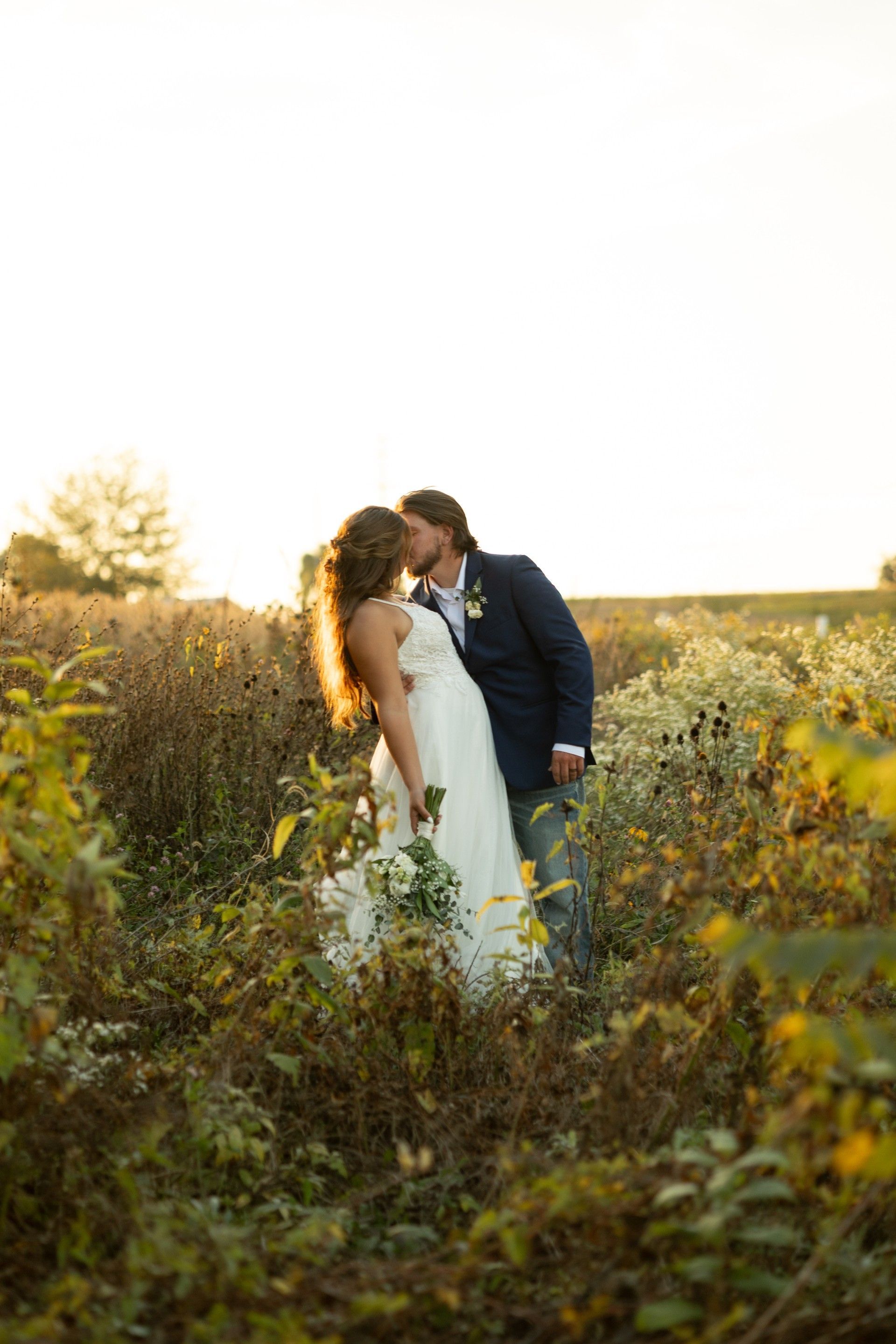 Couple in wedding attire kissing in a sunlit wildflower field at sunset