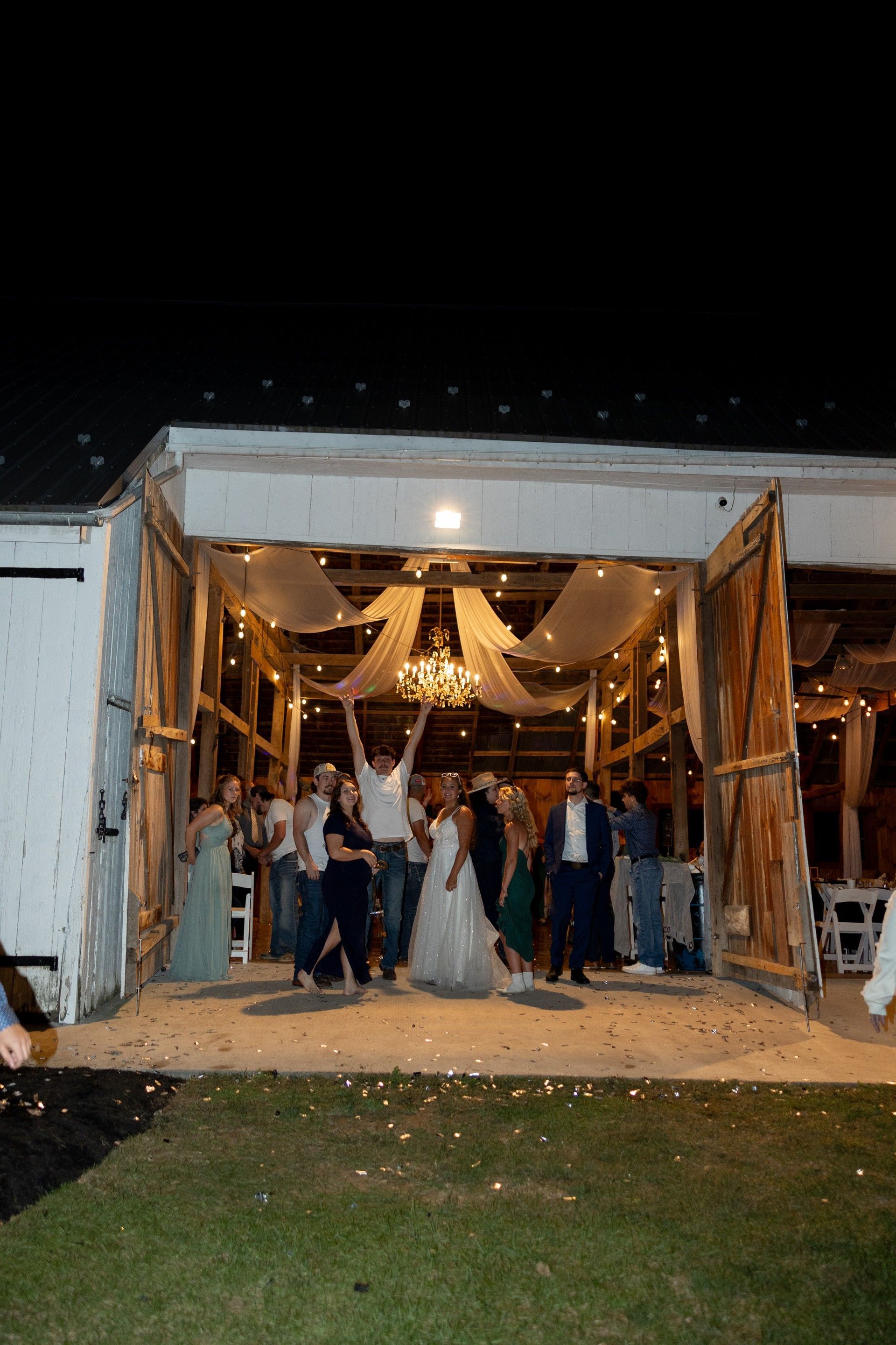 Wedding party gathered in a decorated barn entrance at night, posing under warm lights.