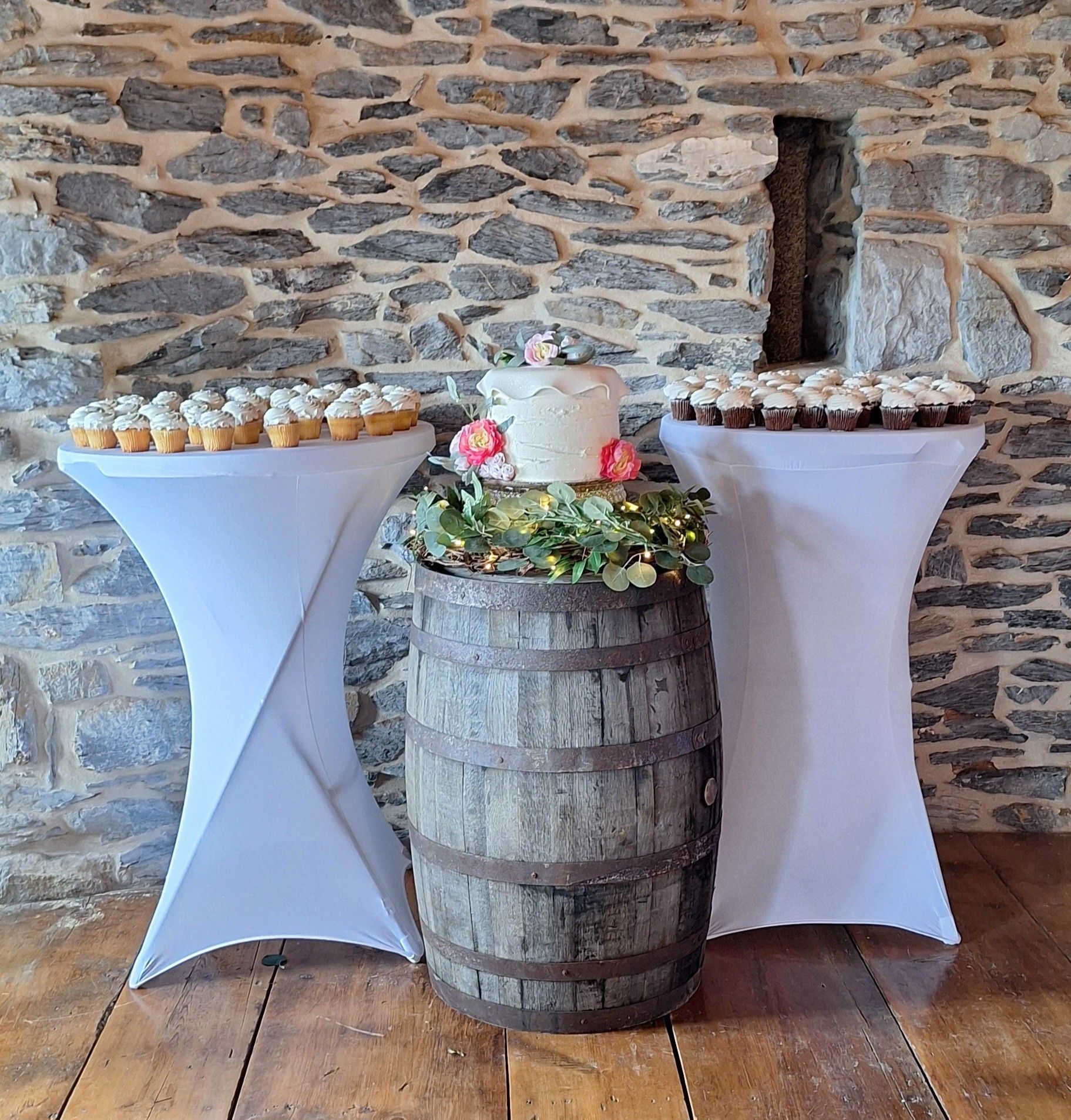 Rustic dessert display with two cocktail tables, cupcakes, and a floral-decorated barrel centerpiece in a stone room