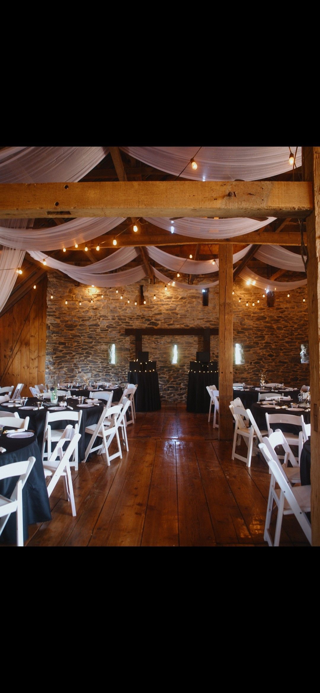 Banquet hall with white chairs, wooden floors, and warm string lights under draped fabric.