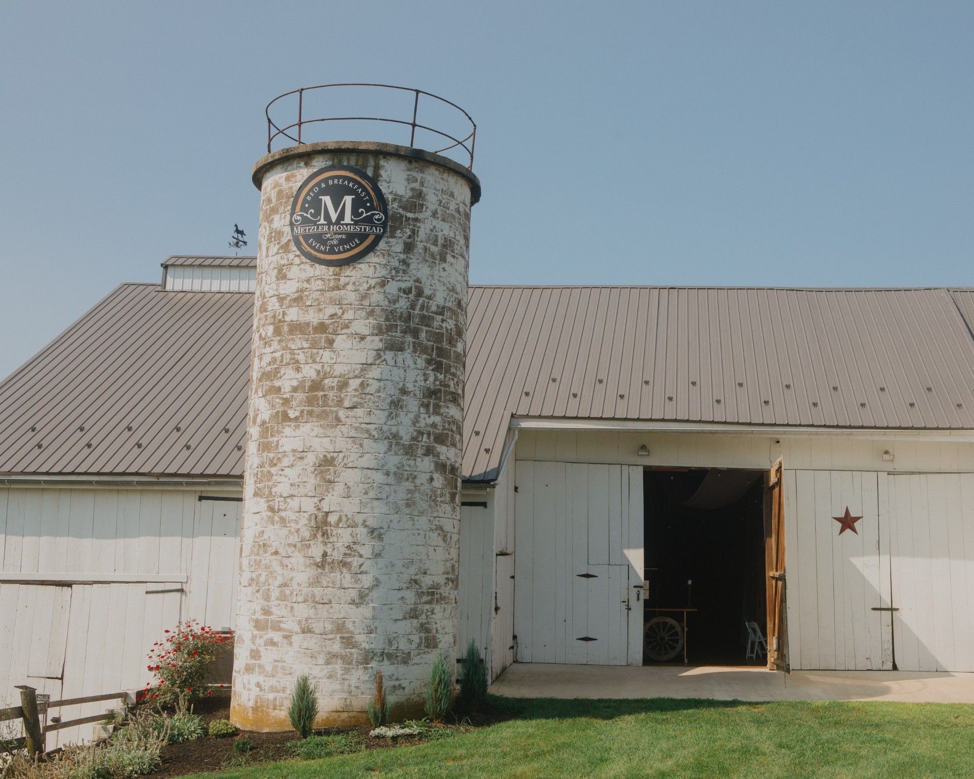 Weathered white barn with a round silo and open white doors under a clear blue sky