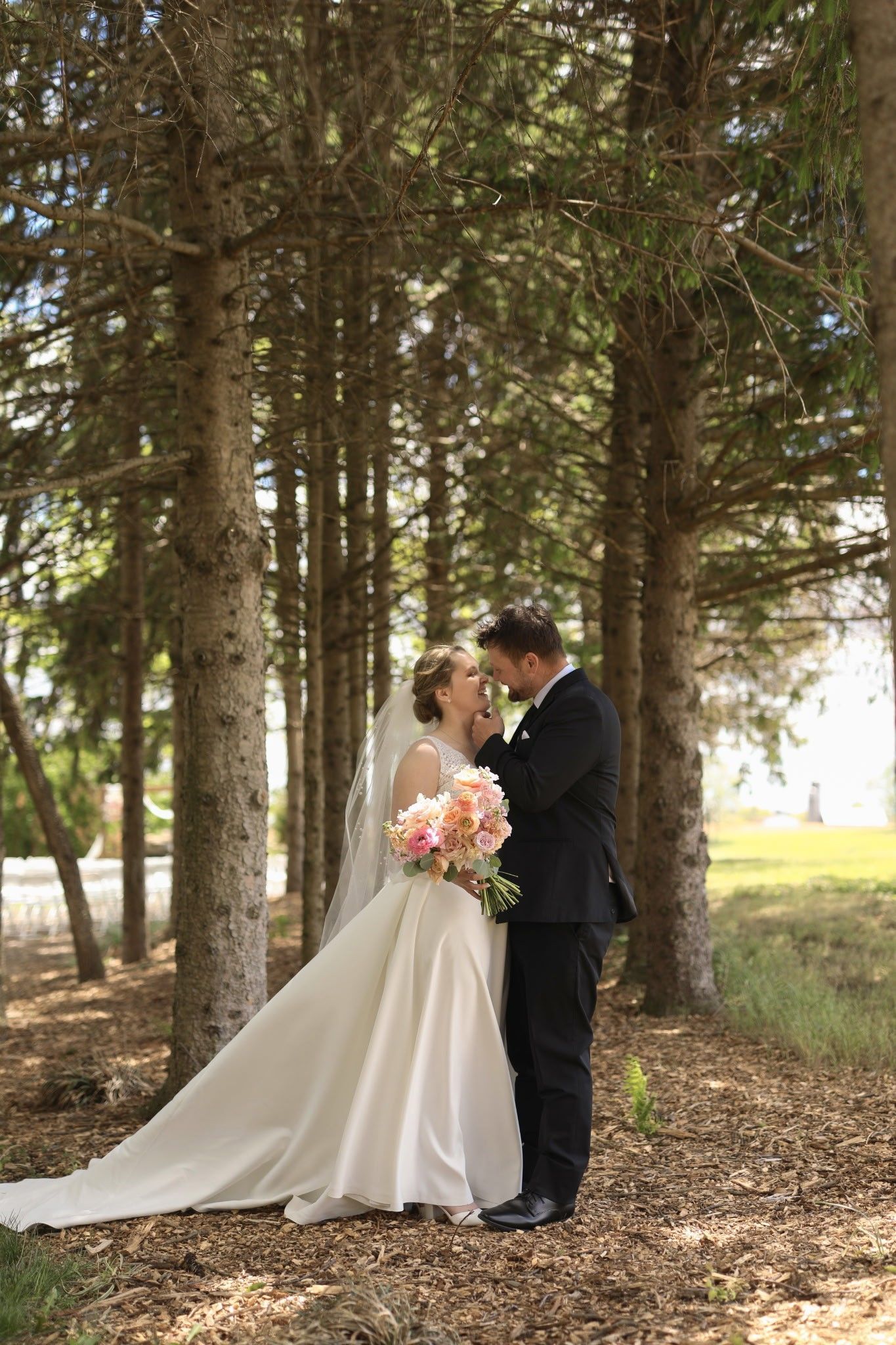Bride and groom embracing in a wooded setting, bride holding a bouquet in a long white gown