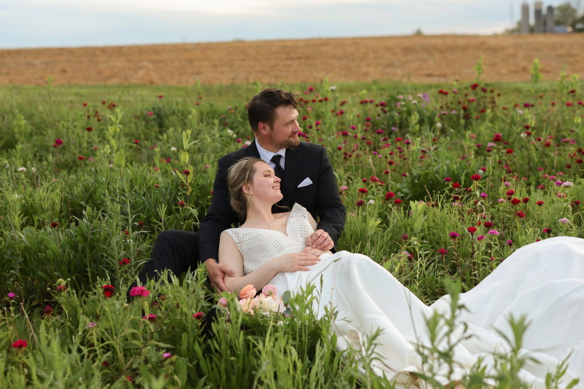 Couple in formal attire relaxing in a wildflower field, with red flowers and a golden horizon.