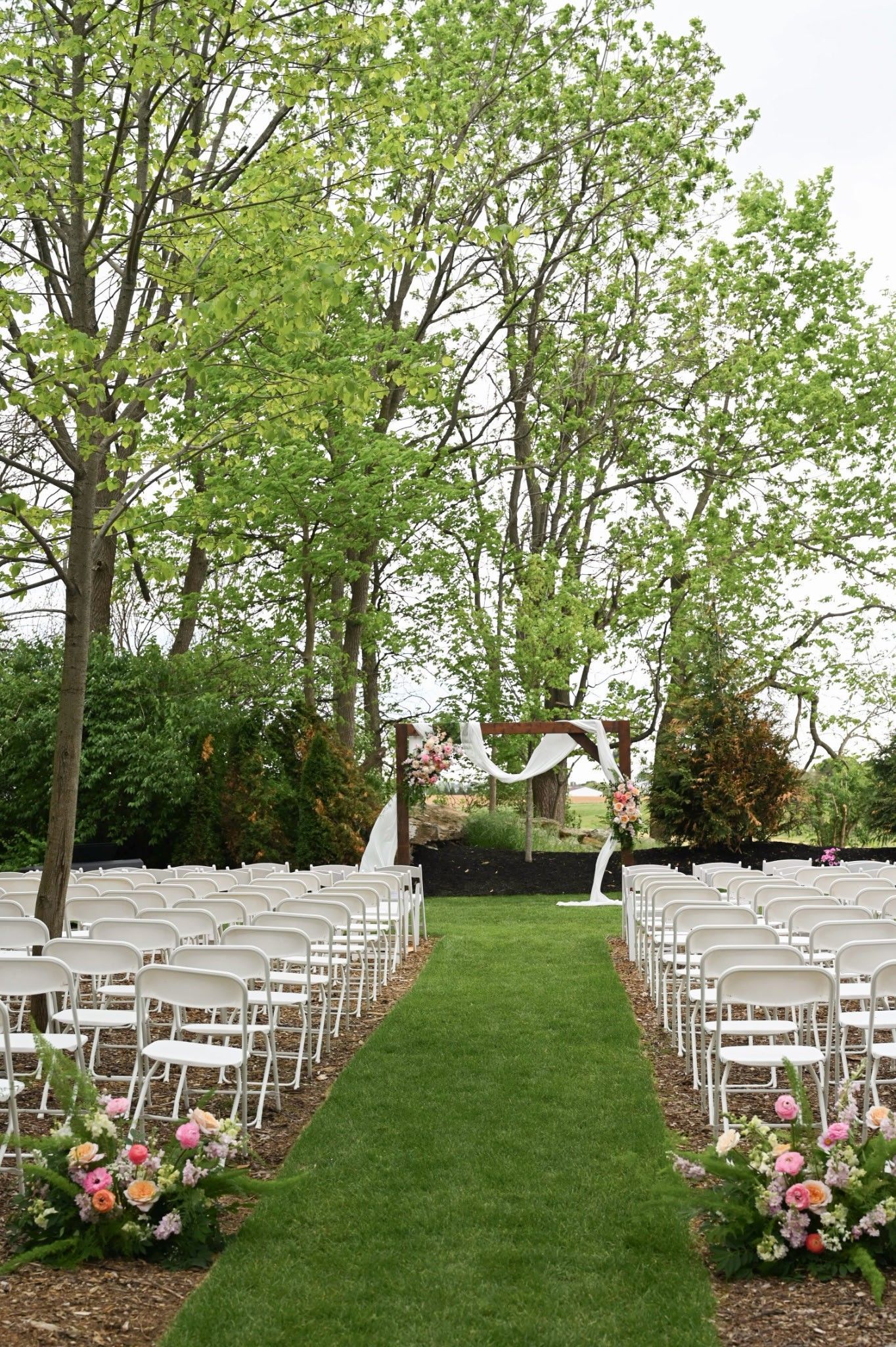 Outdoor wedding aisle with white chairs and pink flowers under tall green trees