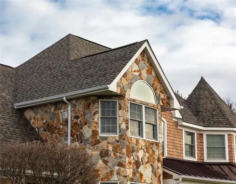 Stone-clad suburban house with gray shingle roof and white-trimmed windows under an overcast sky