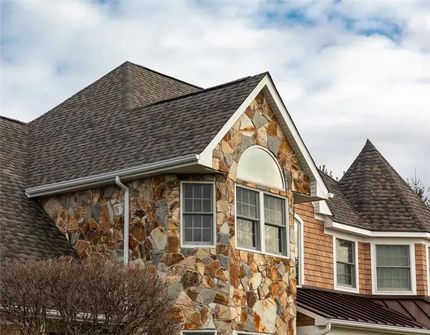 Stone-clad suburban house with gray shingle roof and white-trimmed windows under an overcast sky