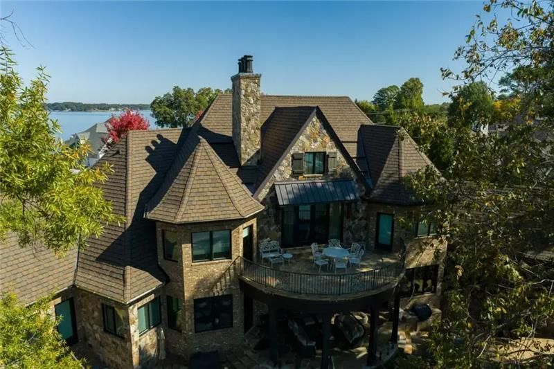 Stone lakeside house with dark shingled roofs, chimneys, and a balcony surrounded by trees and water