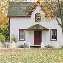 Small white house with red door, green roof, and picket fence surrounded by autumn trees.