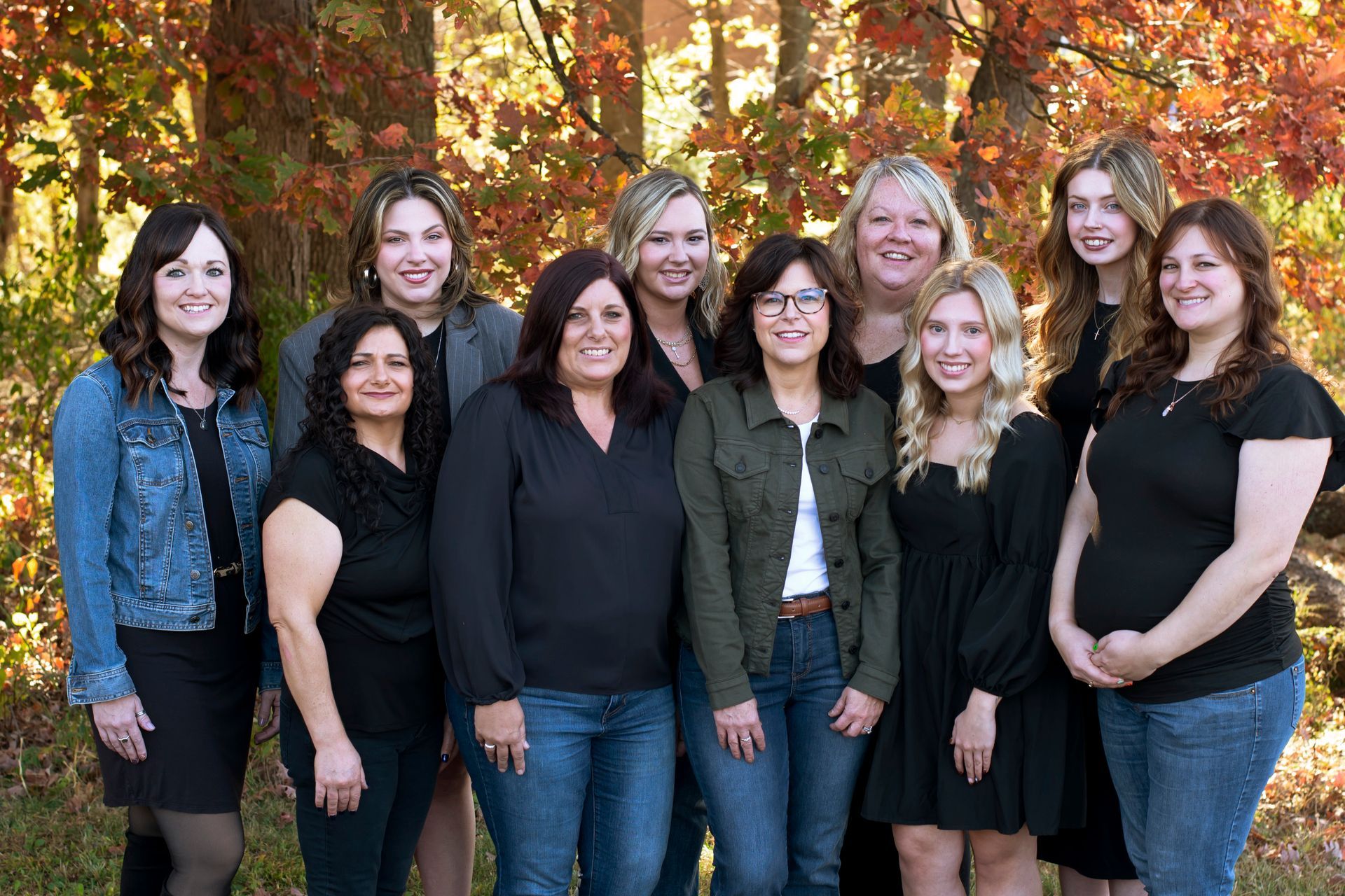Group of 10 women smiling, posing outdoors in front of fall foliage. They wear various casual attire.