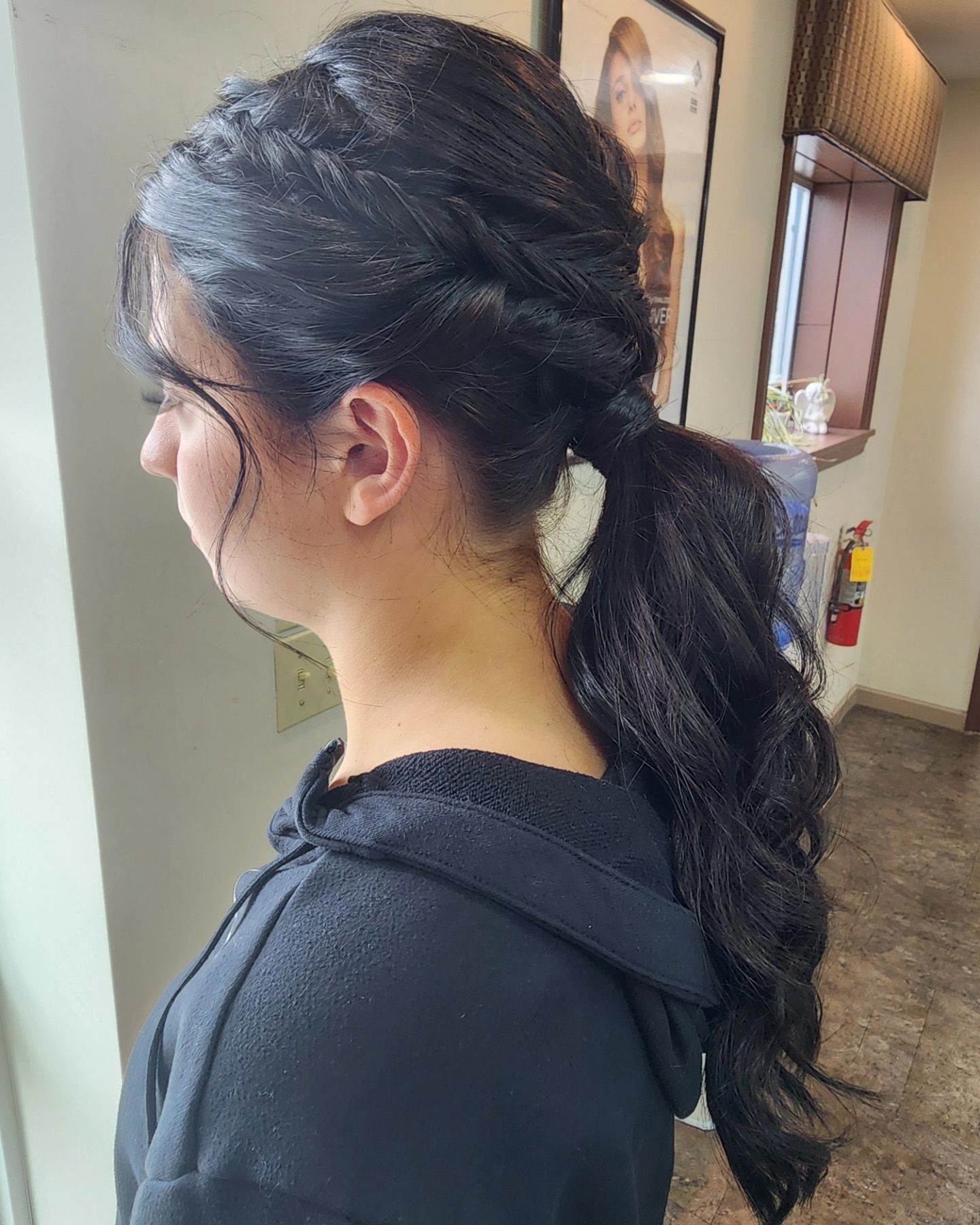 Woman with dark hair in a side ponytail, braided top. Wearing a black hoodie, standing indoors.