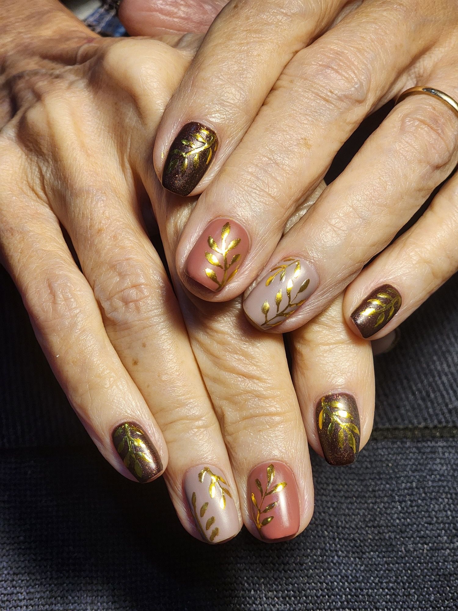 Hands with short, oval-shaped nails. Some are solid brown, others have gold leaf designs on a light pink/beige base.