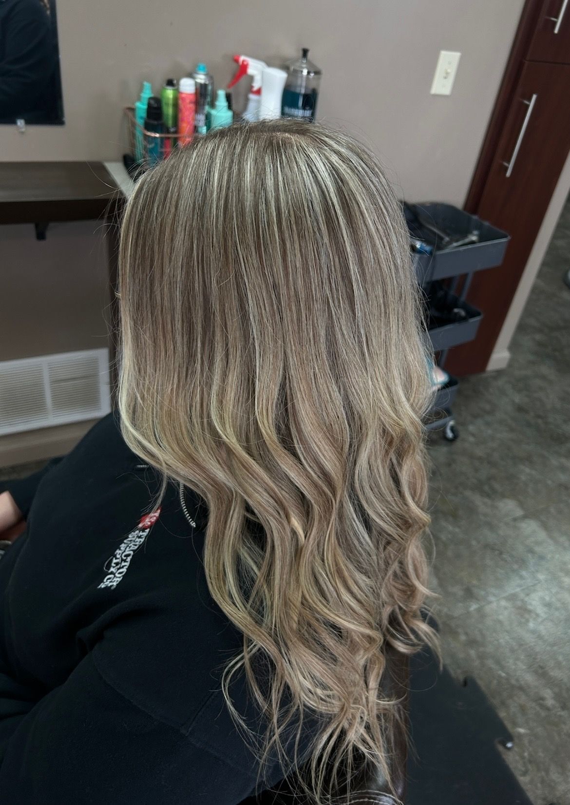 Woman with wavy, blonde-highlighted hair, sitting in a salon chair.