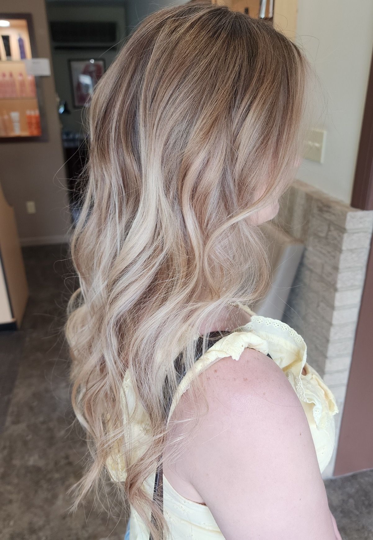Woman with wavy blonde hair, wearing a yellow top, standing near a doorway.