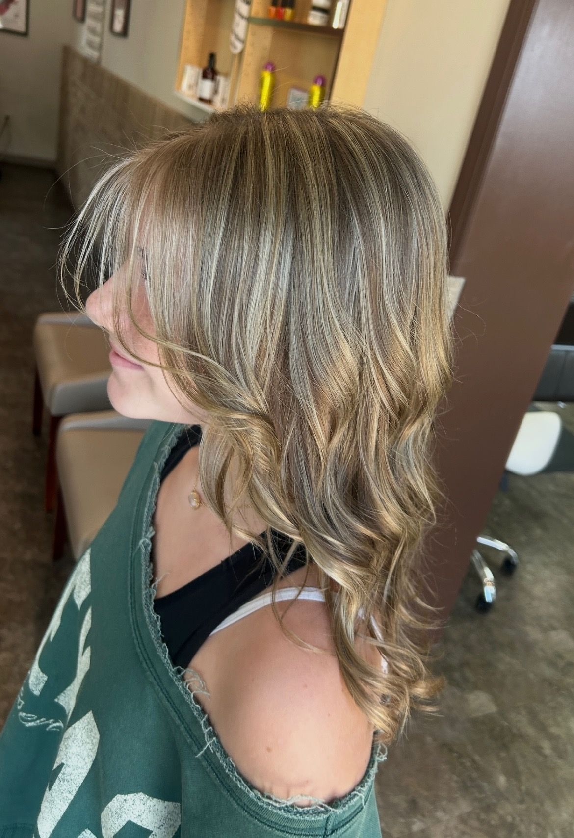 Woman with layered blonde hair at a salon, wearing a green sweatshirt, looking to the side.