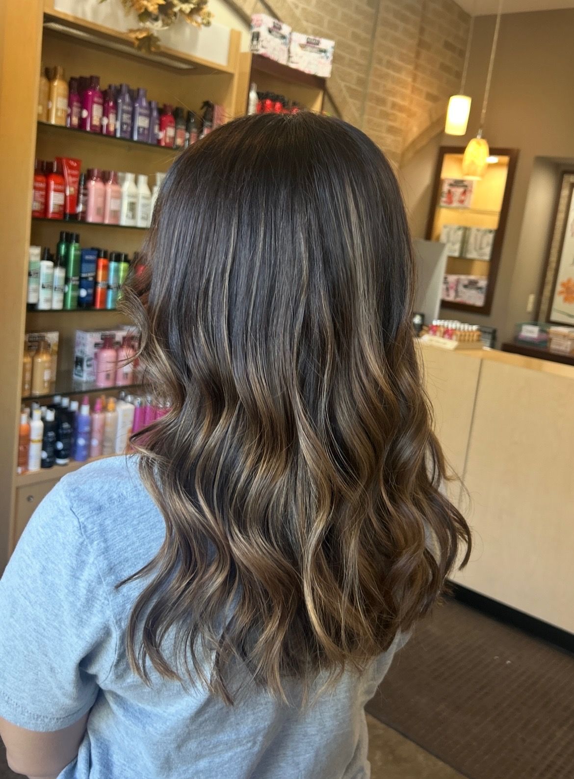 Woman with wavy brown hair, dark roots, light brown highlights, and a blue shirt, in a salon.