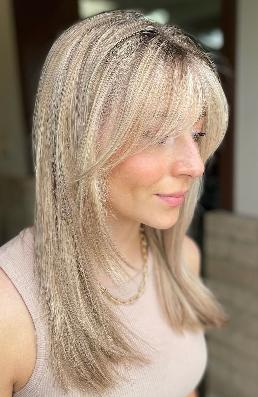 Woman with long blonde hair, side bangs, wearing a pink tank top, gold necklace, looking right, neutral expression.