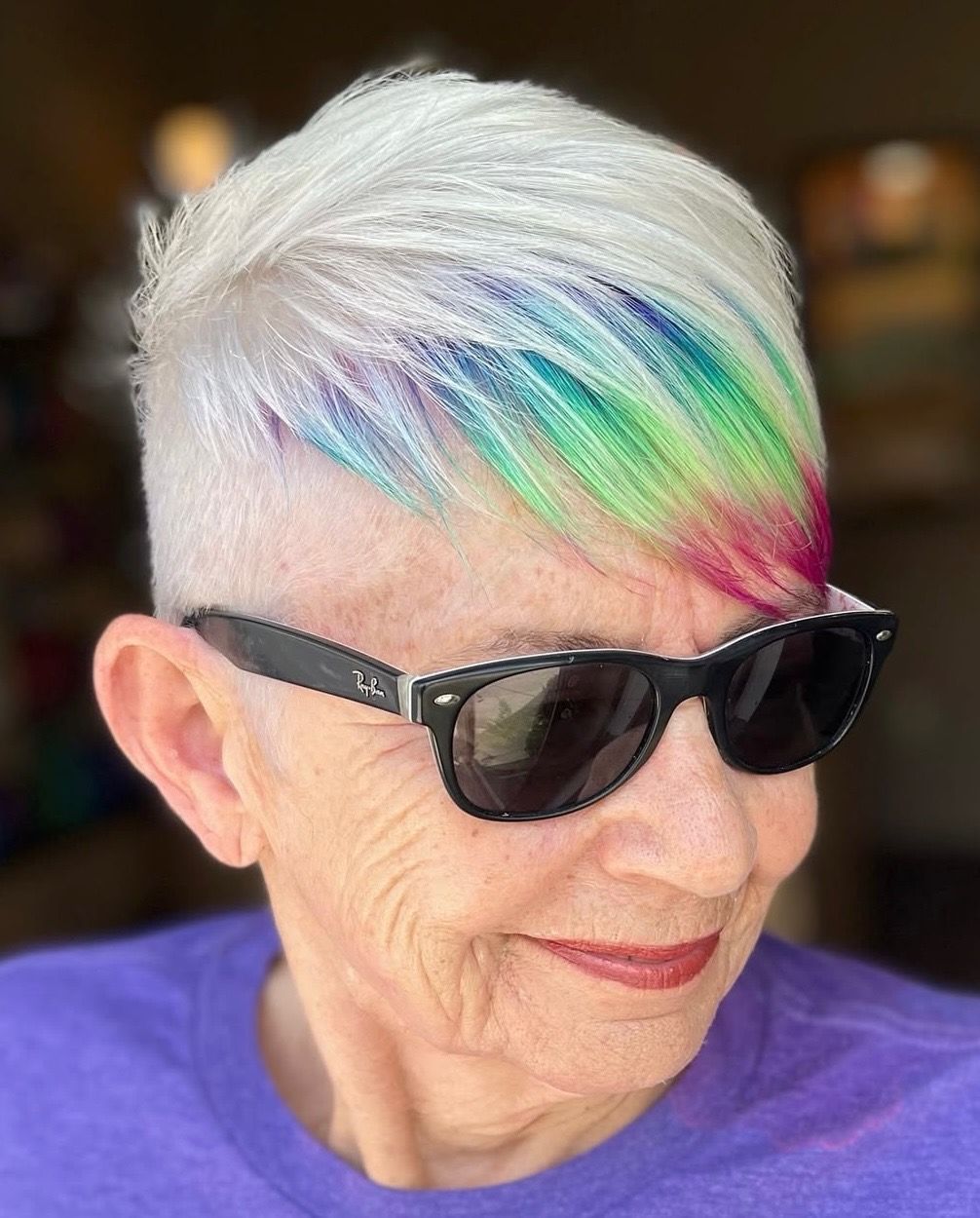 Woman with short, white hair with rainbow highlights wearing sunglasses and a purple shirt.