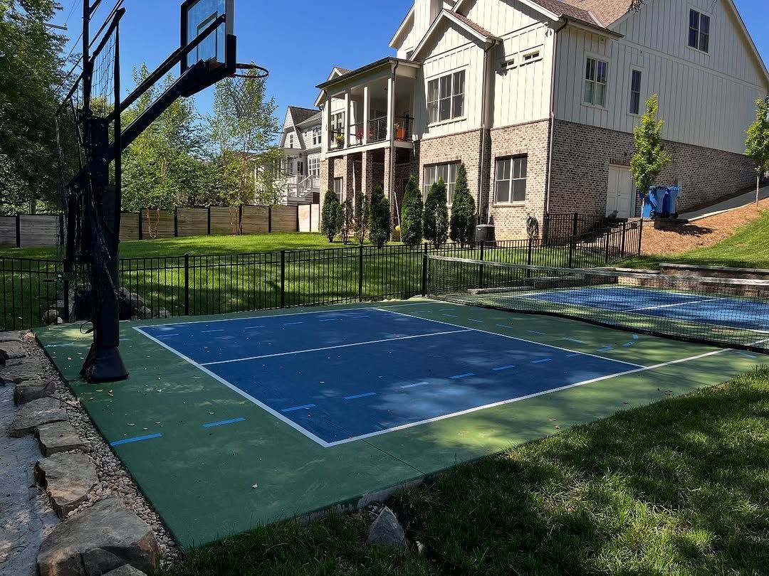Basketball court in a backyard with a house in the background. Blue court with a black hoop.