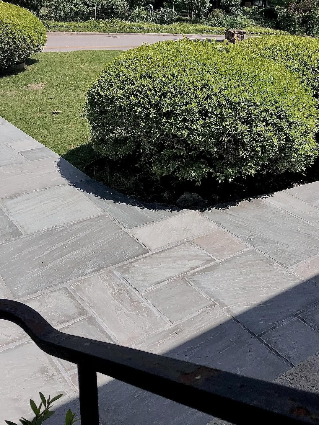 Stone path leading to a manicured garden with a large green bush and a black railing.