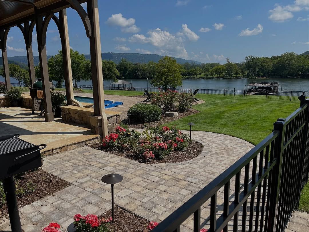 Patio overlooking a river, with flowers, grill, and gazebo-style structure. Sunny day, blue sky, and mountains in the distance.
