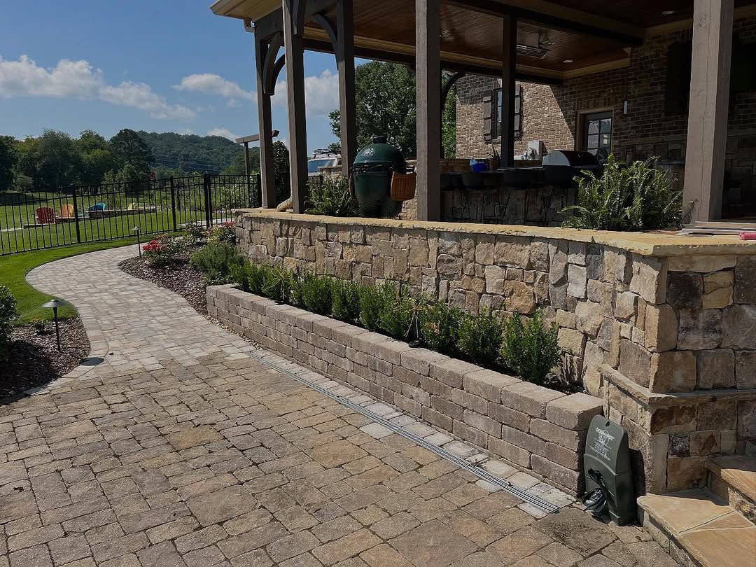 Stone patio and pathway leading to a covered outdoor kitchen. Green shrubs line a stone wall.