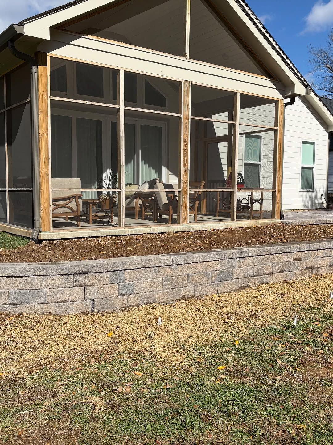 Screened-in porch attached to a house with a stone retaining wall and a lawn in the foreground.