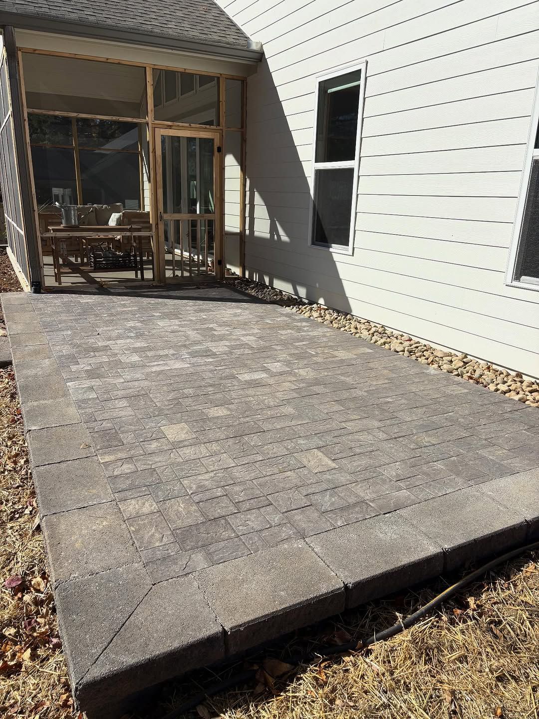 Paver patio next to a house with a screened porch, surrounded by grass and small rocks.