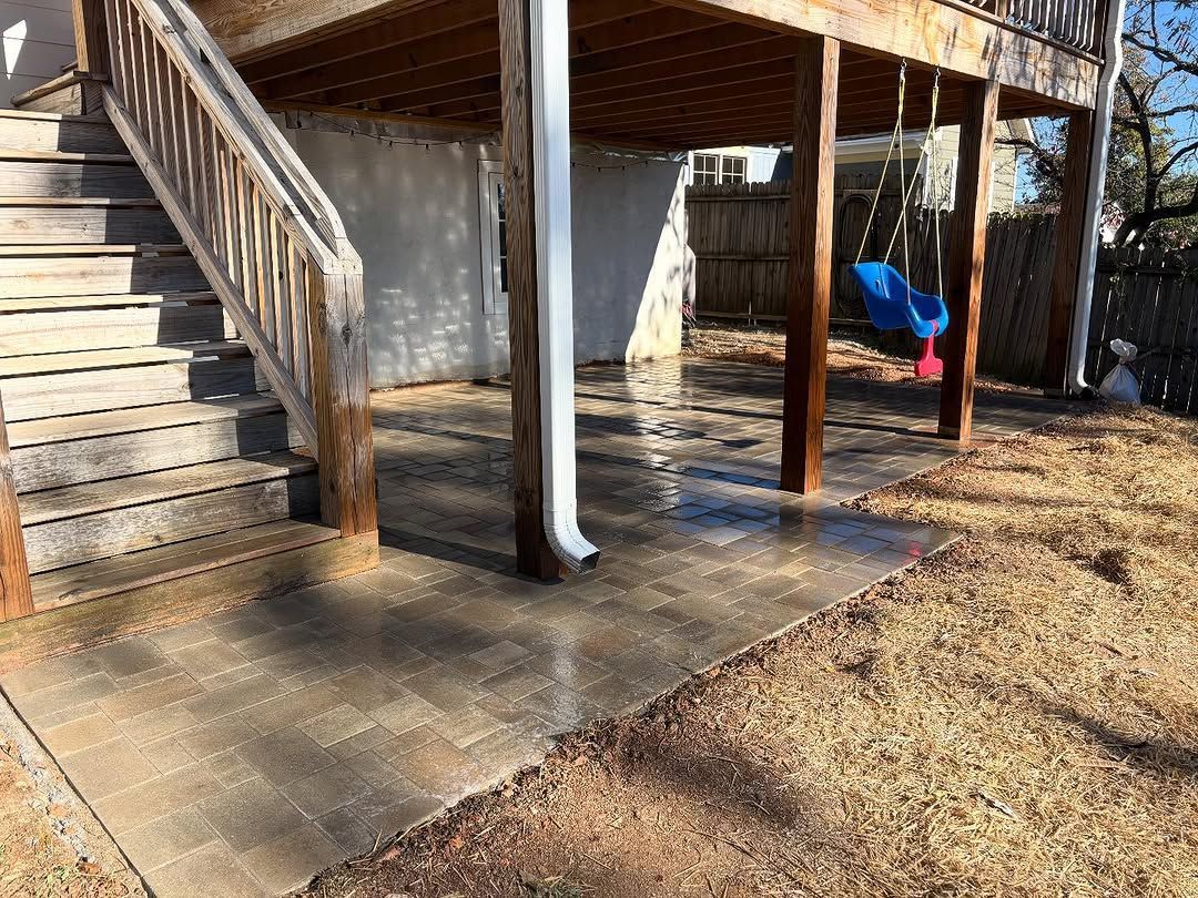 Concrete patio under a wooden deck with stairs, a blue swing, and a gutter, on dry grass.