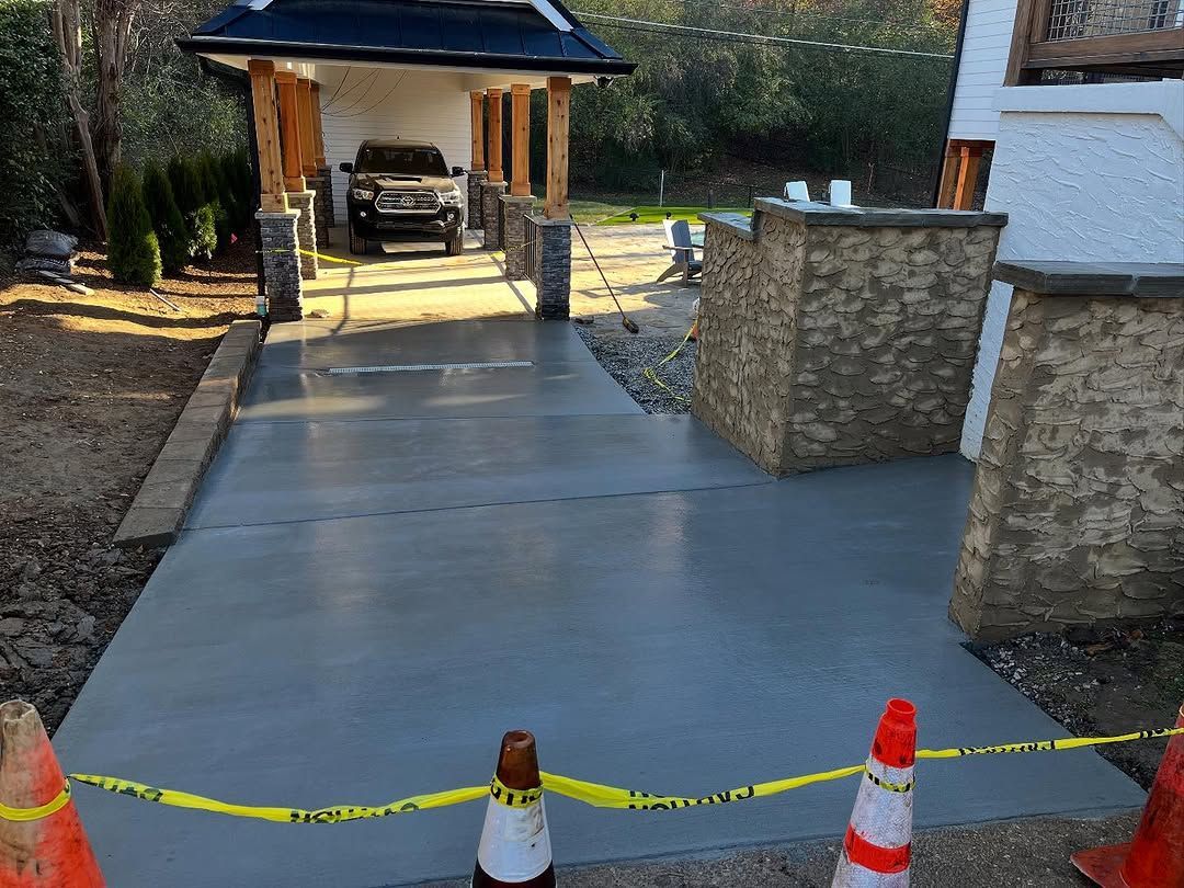 Newly poured concrete driveway with a vehicle parked in a wooden carport; orange cones and caution tape.