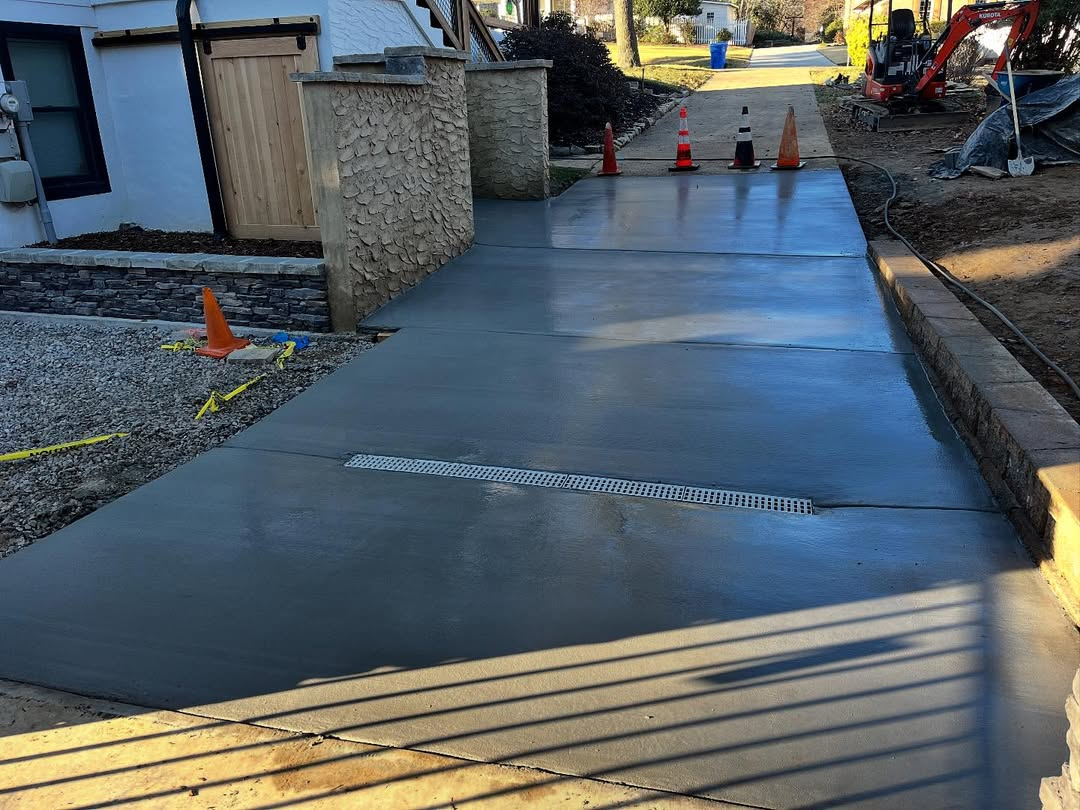 New concrete driveway with expansion joints, sidewalk construction. Construction cones, columns, excavator.