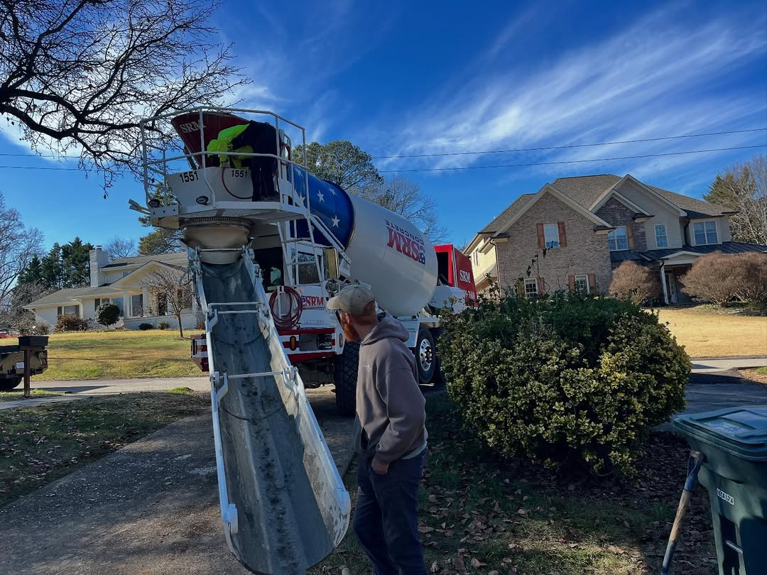 Cement truck pouring concrete on a driveway; worker in foreground, residential setting with blue sky.