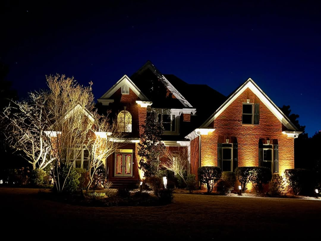 A brick house illuminated by outdoor lighting at night, with trees and shrubs in the front yard.
