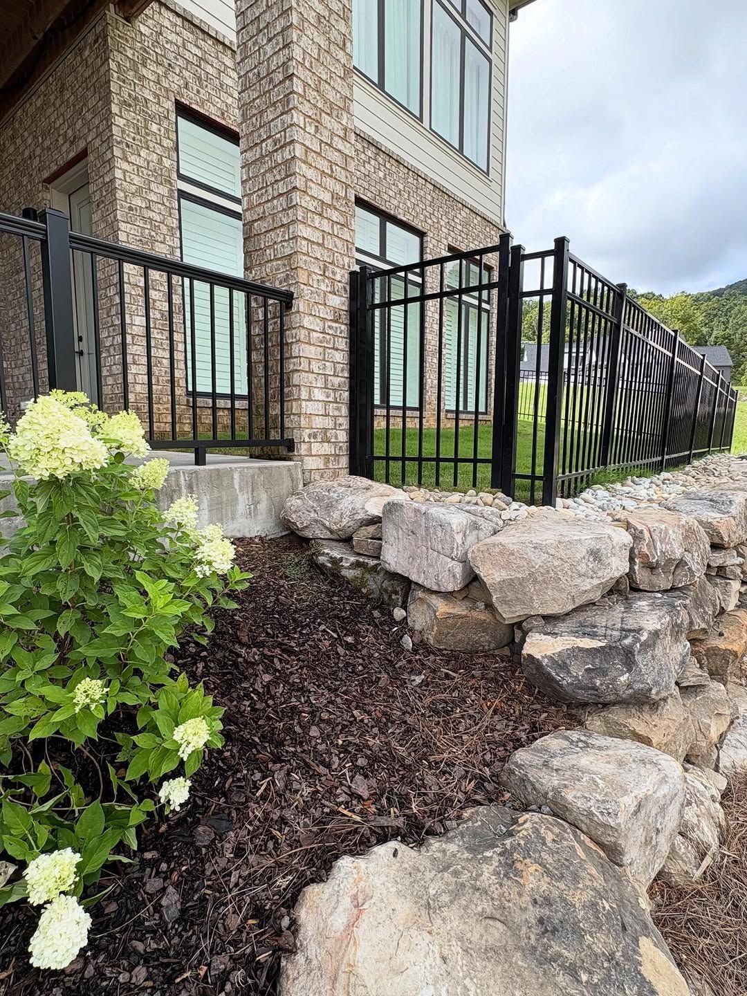 Black metal fence on a stone wall. A building with large windows is in the background. Hydrangeas in the foreground.