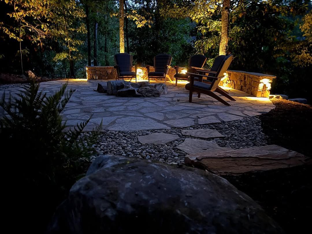 Outdoor patio with seating around a fire pit, lit by warm lights in a wooded setting.