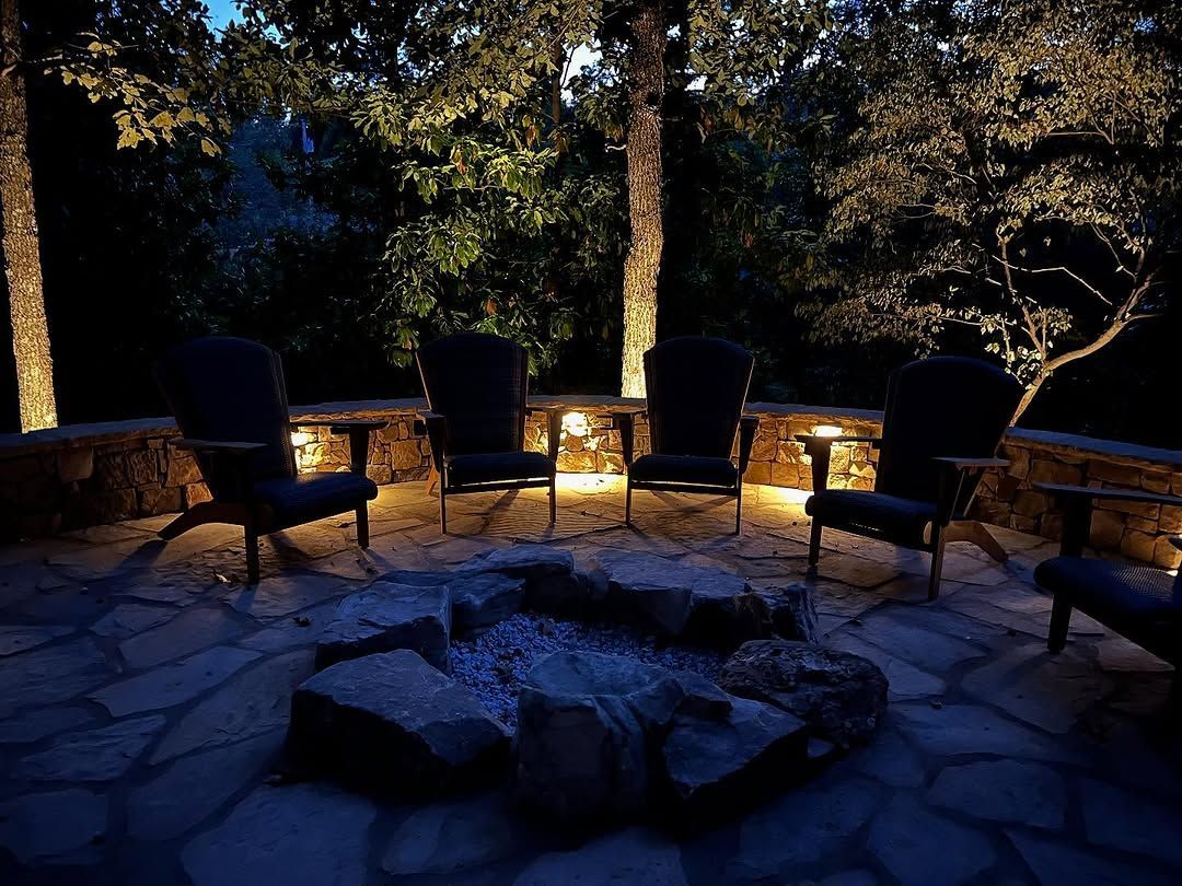Outdoor patio at night with illuminated trees, chairs around a fire pit, and stone pathway.
