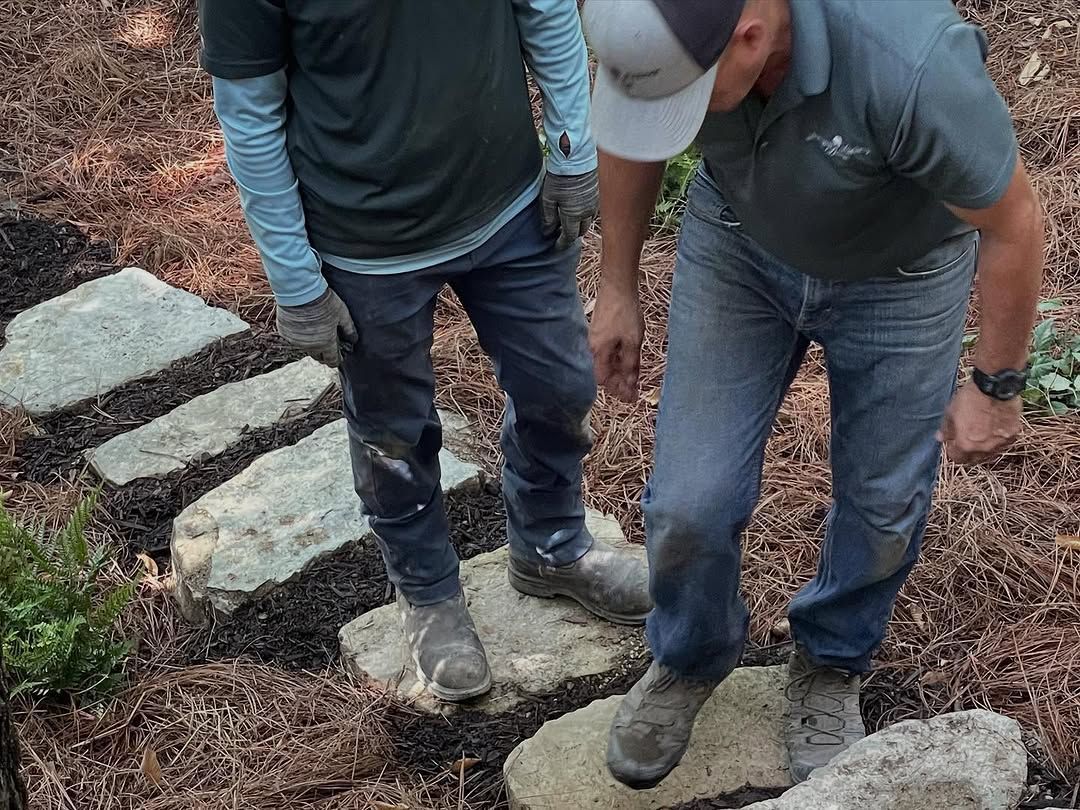 Two people walking on stone steps in a garden bed. One man wears a hat and the other has a long-sleeved shirt.