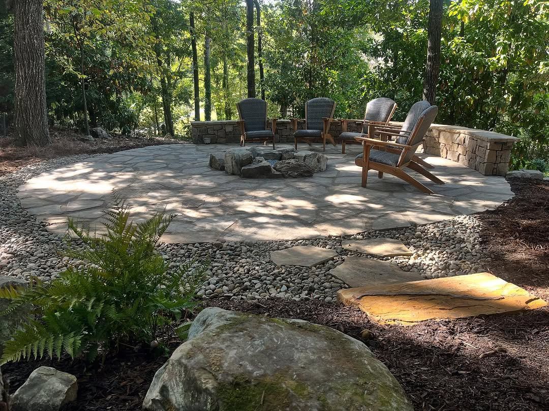 Stone patio with fire pit, seating, and surrounding greenery in a wooded area.