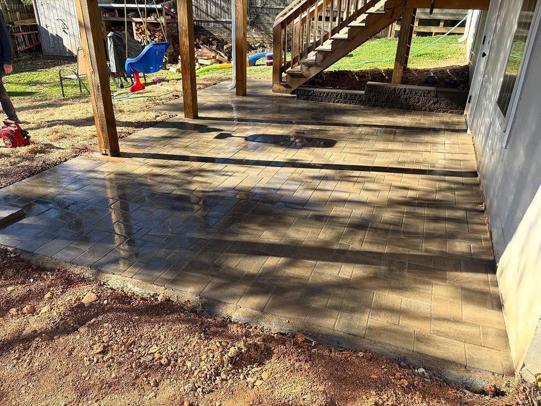 Newly installed brick patio under wooden deck; wet surface, brown soil surrounds.