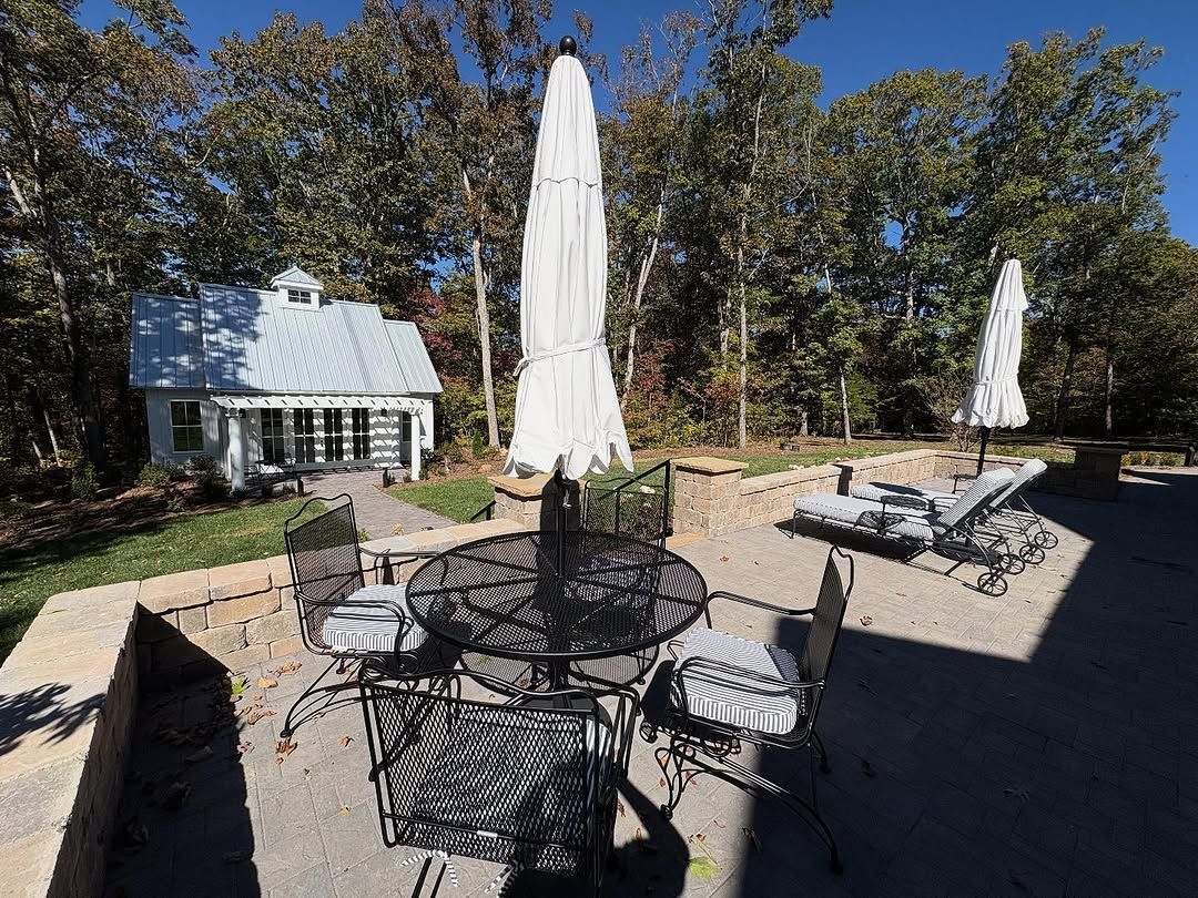 Patio with table, chairs, umbrellas, lounge chairs, and small white shed in a wooded setting.