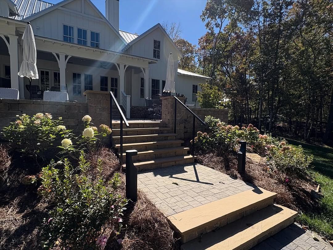 Stone steps leading up to a white farmhouse with a black railing, surrounded by bushes and trees.