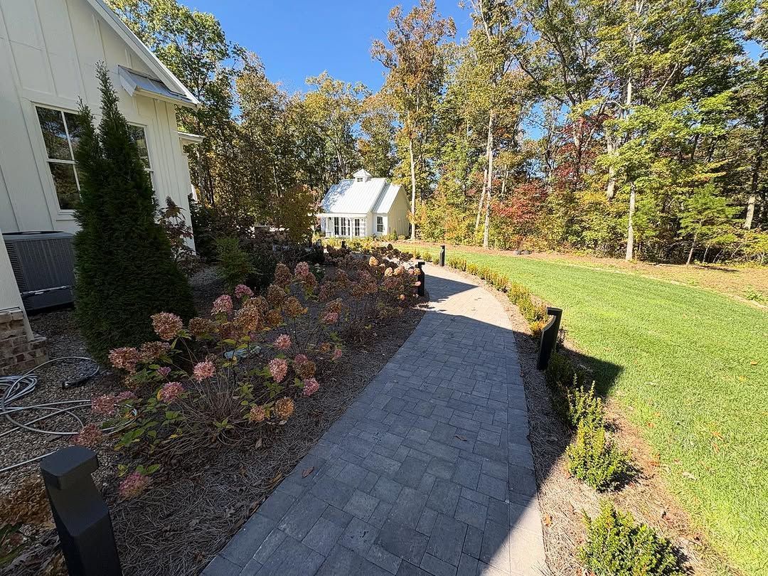 Brick path winds through a garden, past landscaping, towards a white building and trees under a blue sky.