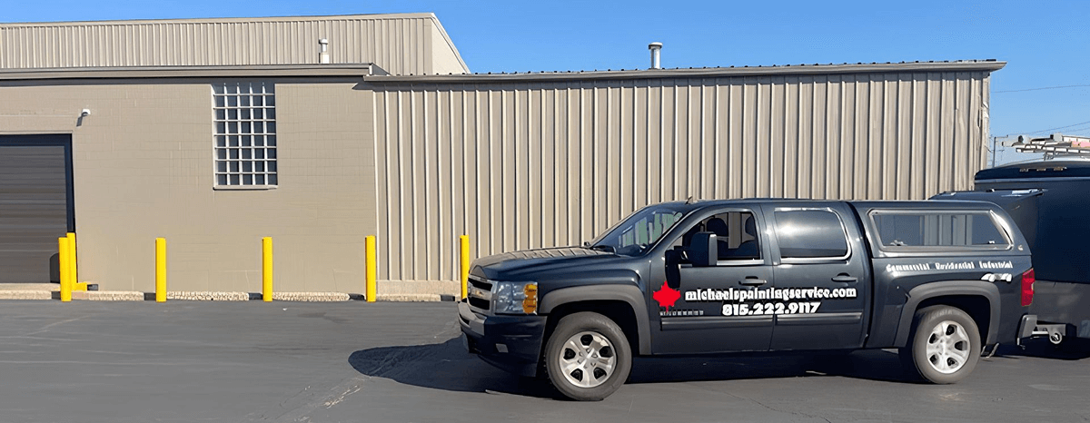 Beige metal warehouse exterior with roll-up door and yellow bollards on an asphalt lot.