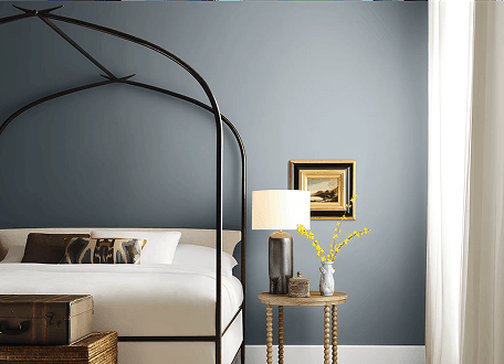 Blue bedroom with black canopy bed, bedside lamp, and small table with flowers by a window