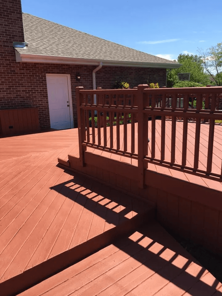 Red wooden deck with railing beside a brick house and white door in bright sunlight