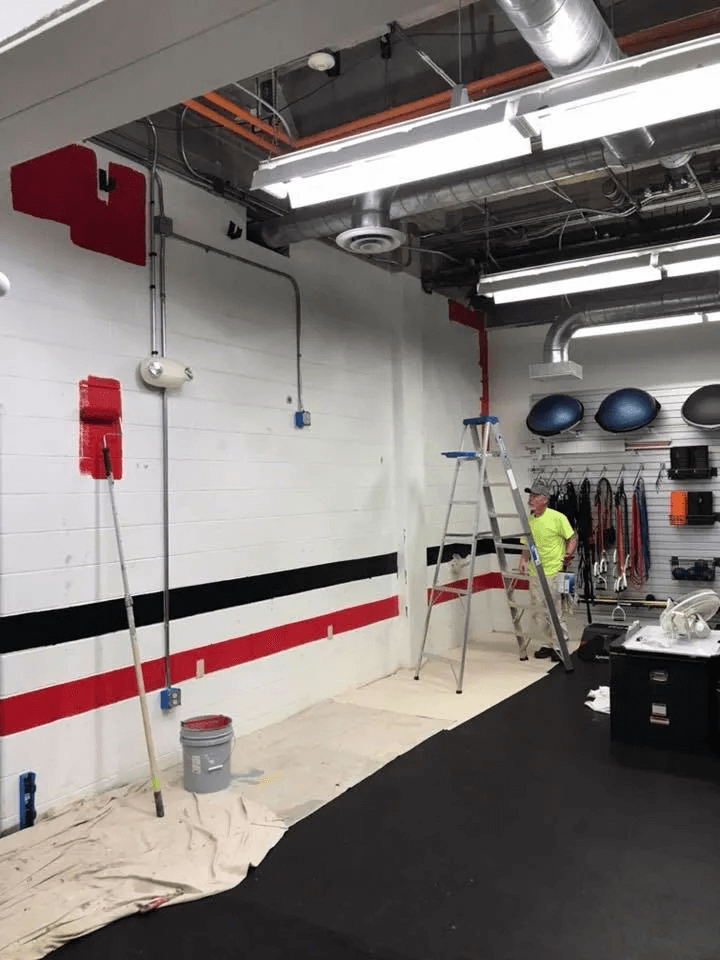 Worker on ladder in industrial garage, painting white wall with red and black stripes.