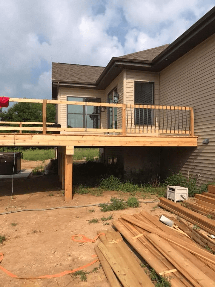 Wooden deck under construction beside a house, with stacked lumber and a partly cloudy sky.