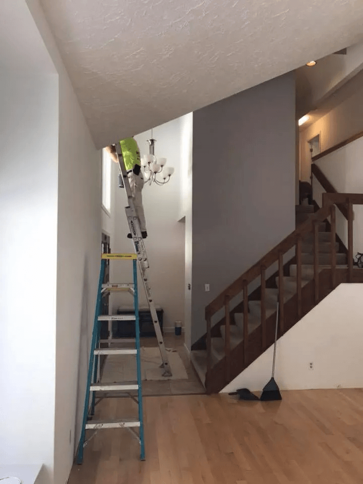 Worker on a ladder painting a hallway ceiling beside a staircase.
